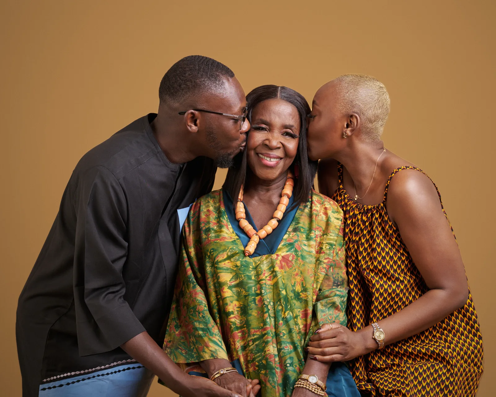A son and daughter kiss their smiling mother on both cheeks, she wears a green batik kaftan and coral beads