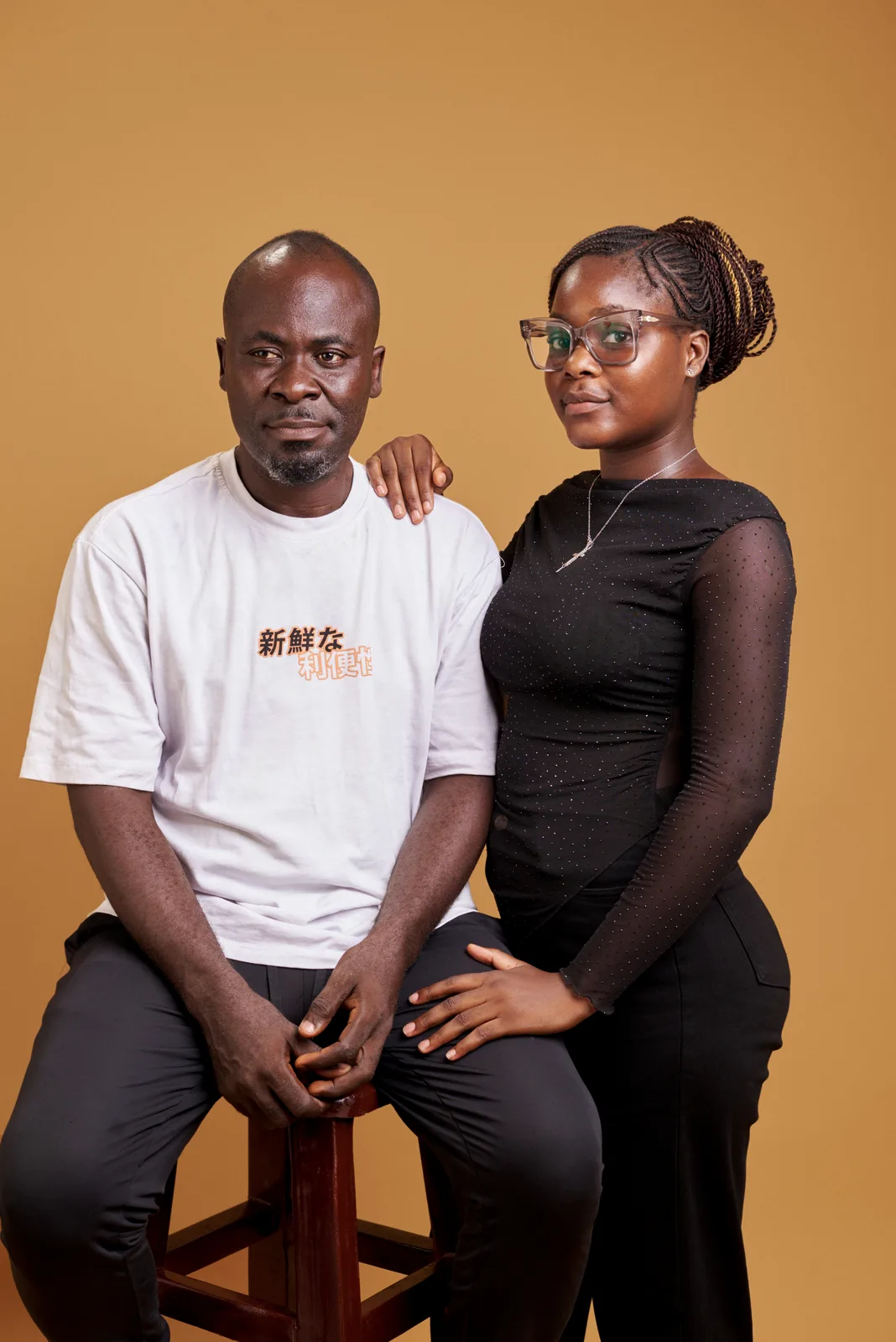 A father in a white T-shirt seated on a wooden stool, his daughter standing beside him with a hand on his shoulder