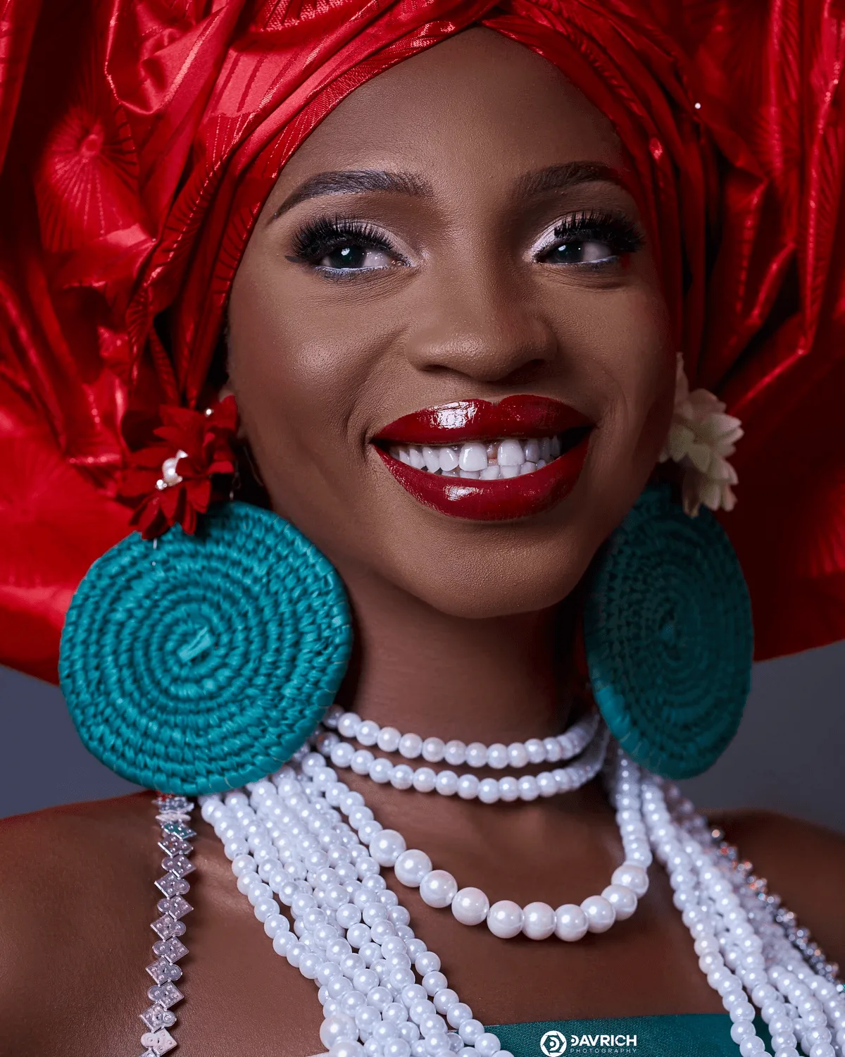 Smiling woman in a red gele with teal woven earrings and layered pearl necklaces