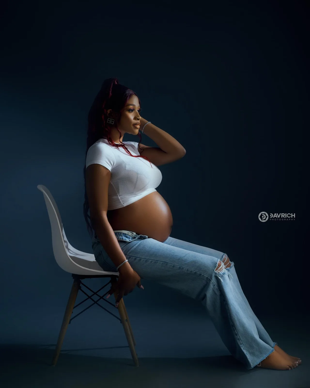 Maternity portrait of an expectant mother seated in a white crop top and jeans against deep blue