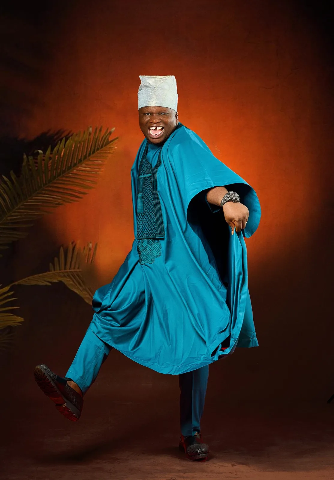 Man in a teal agbada and white cap dancing against a warm orange backdrop
