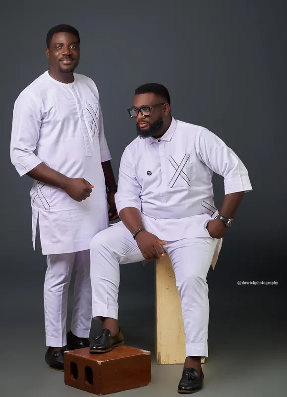Two men in matching white native attire, one standing and one seated, in the studio