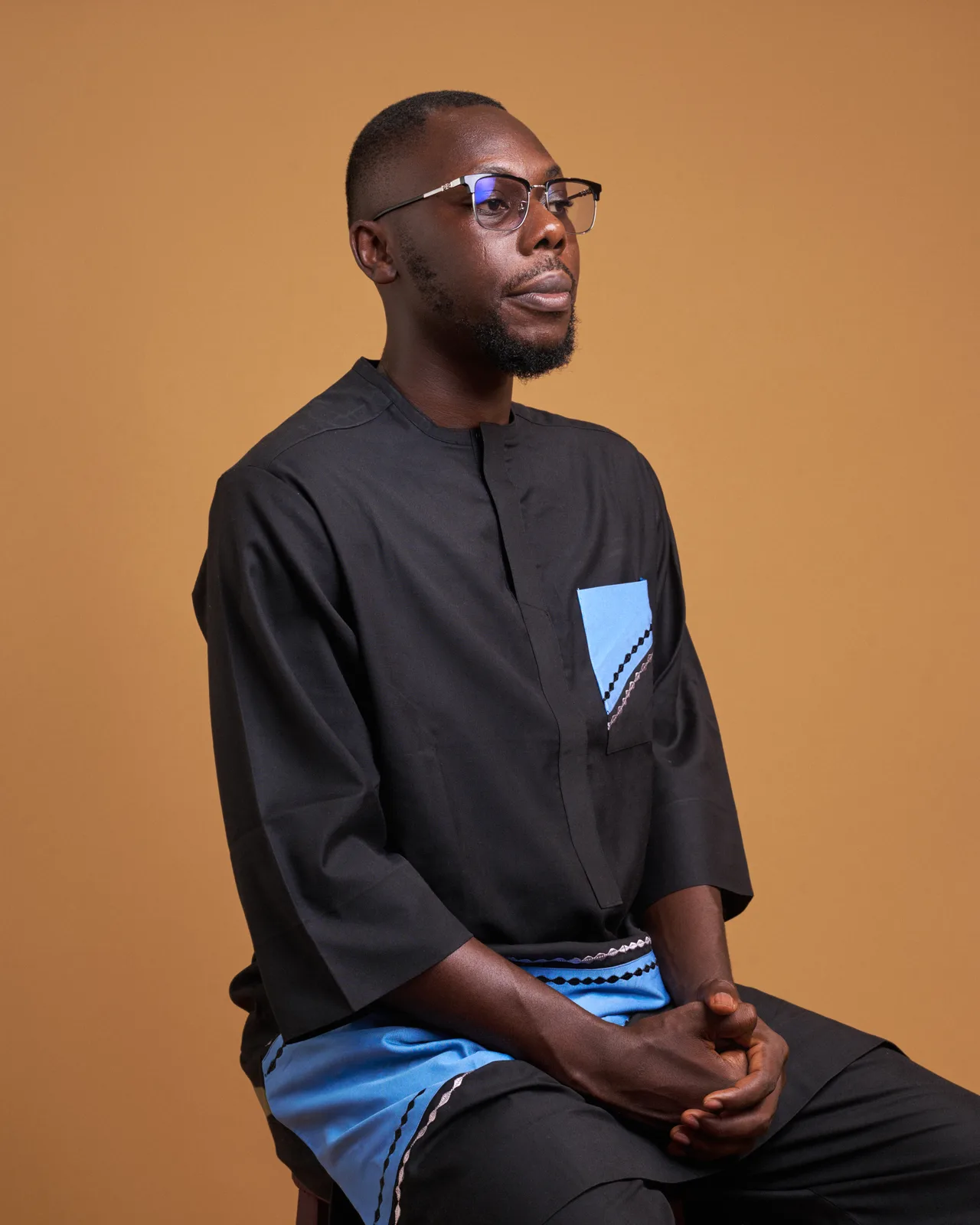 Man in glasses and a black and blue kaftan, seated with hands clasped, looking away against a warm caramel backdrop